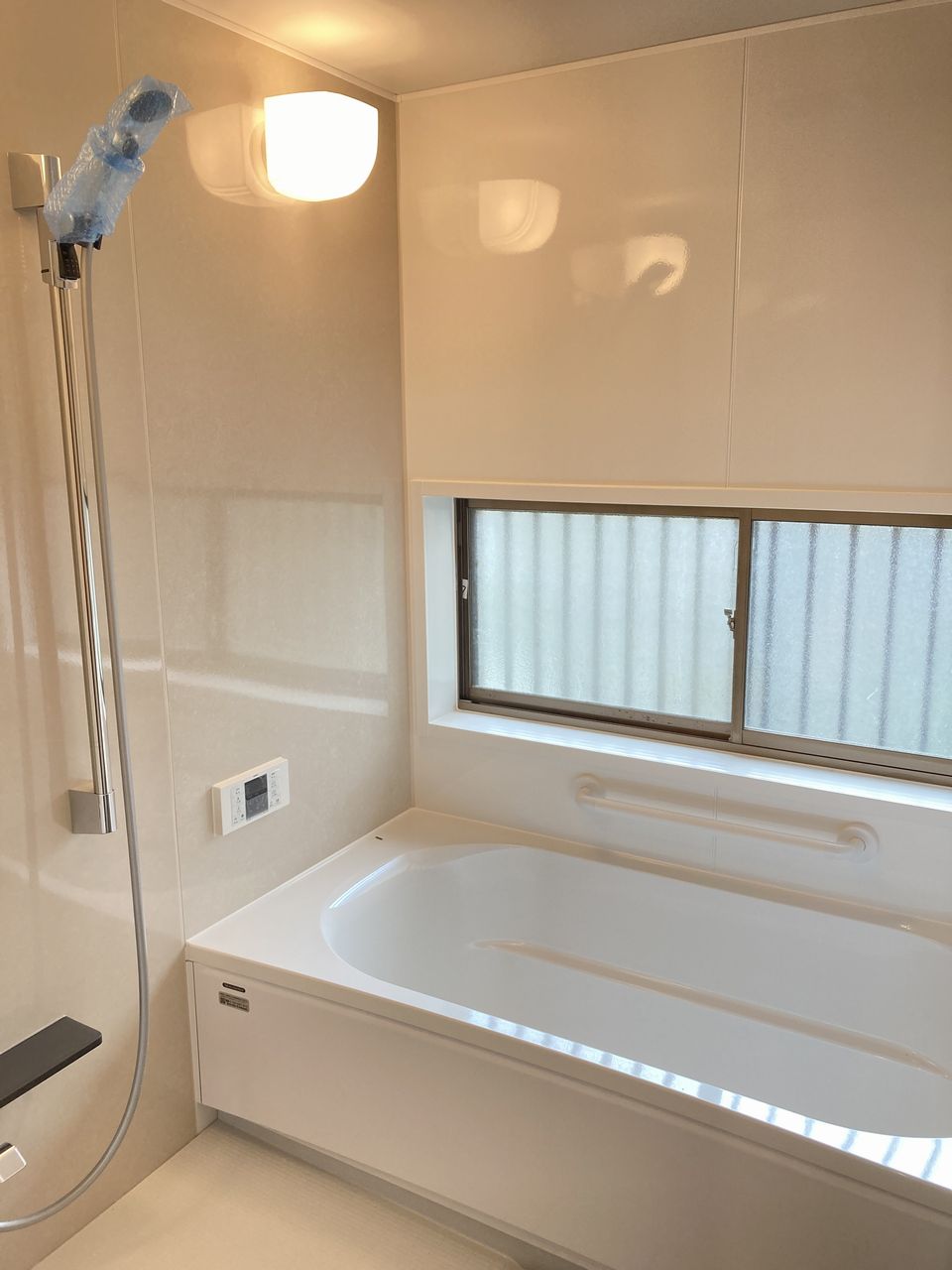 浴室の写真。浴槽の横に手すりがあり、壁面にシャワーと操作パネル、窓から自然光が入る清潔感のある浴室です。