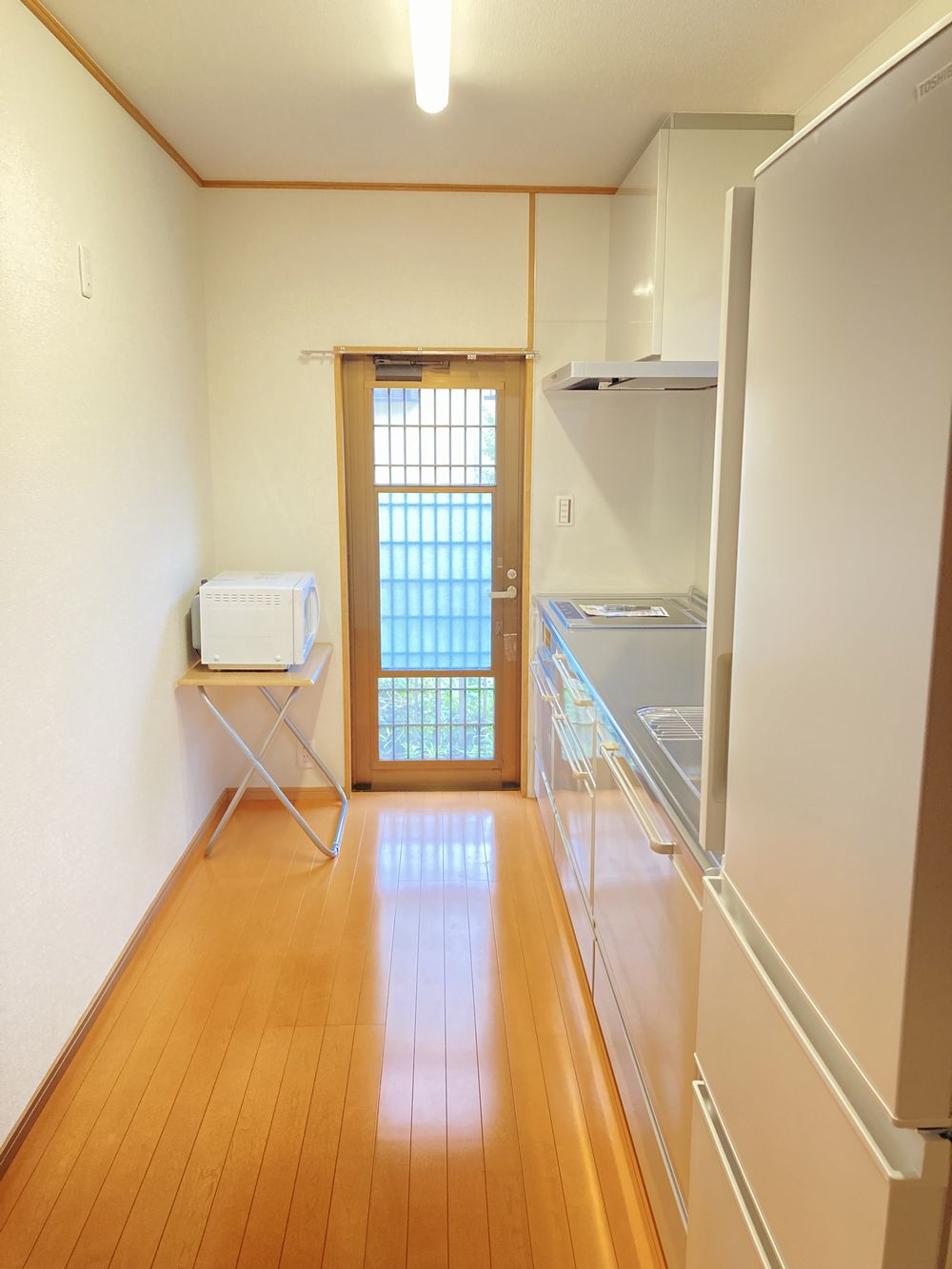 キッチンの写真。右側にシステムキッチンと冷蔵庫が並び、奥に勝手口のドアがある細長い動線の明るい空間です。