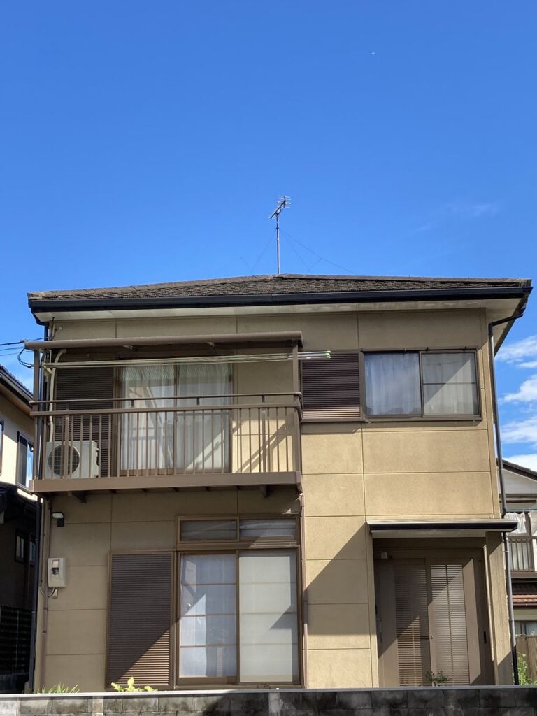 快晴の空のもと、玄関側から見た２階建てのグループホームムティ。クリーム色の外壁で、住宅街に馴染んだ一戸建て風の建物。右手側に玄関がある。