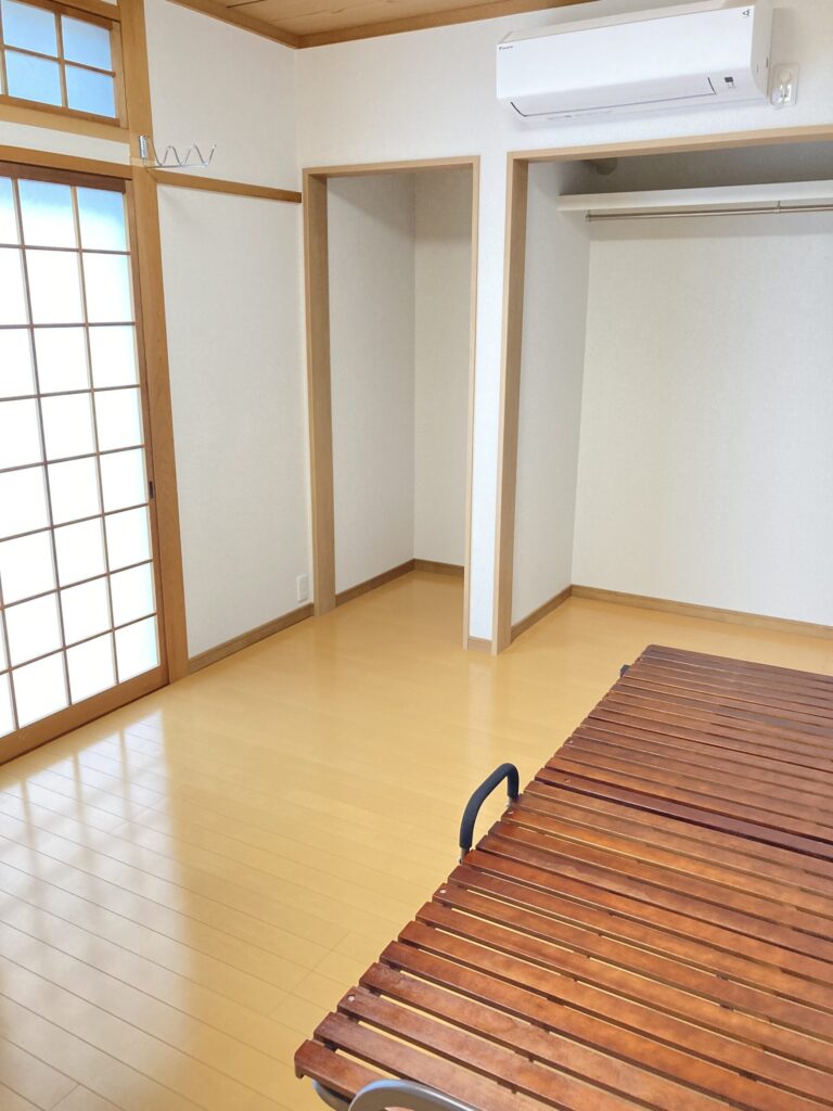 木目調フローリングと白い壁紙の個室。左に日当たりの良い窓と部屋干し用フック、右に折りたたみベッドが置かれている。